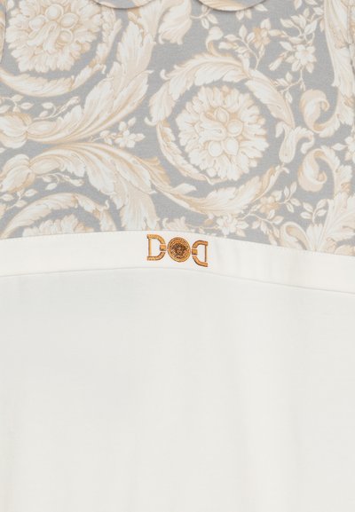 Blomstrete topp i lys beige og grå med et hvitt stoffpanel. Har en subtil logo i gulltråd på brystet.