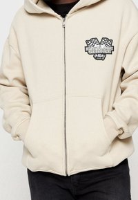 Sweat à capuche beige avec fermeture éclair, fabriqué en tissu doux, avec un motif graphique frontal représentant des drapeaux à damier et le texte "Fastlane Service". Poches avant incluses.