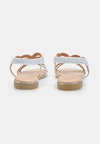 Sandales argentées avec des brides texturées et un design à bords festonnés, dotées d'une semelle plate et d'un dos ouvert pour un enfilage facile.