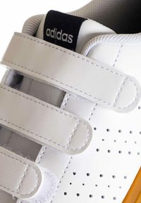 Zapatilla blanca con correas de Velcro, diseño perforado, acentos de costura y una suela de goma marrón. Presenta un logo negro en la lengua.