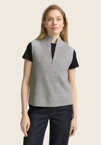 Graue Strickweste mit hohem Kragen und Reißverschluss, mit geripptem Saum. Trägt man über einem schwarzen T-Shirt und kombiniert mit dunklen Jeans.