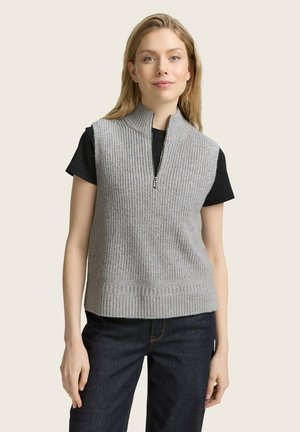 Gilet en tricot gris avec un col montant et une fermeture éclair, doté d'un ourlet côtelé. Porté sur un t-shirt noir et associé à un jean foncé.