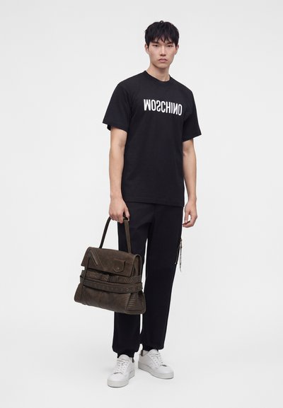 Νέος άνδρας όρθιος, φοράει μαύρο μπλουζάκι Moschino, μαύρο παντελόνι, λευκά αθλητικά παπούτσια, κρατώντας μια μεγάλη καφέ δερμάτινη τσάντα στο δεξί του χέρι.