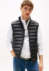 Jovem com cabelo encaracolado vestindo um colete acolchoado preto por cima de um pulôver branco e jeans azuis, em pé com uma mão no bolso.