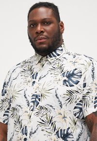 Camisa de manga corta de tela ligera, con un fondo blanco y patrones tropicales de flores y hojas en verde y azul.