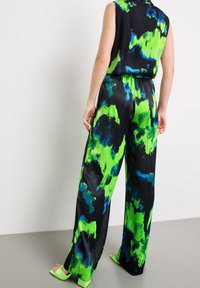 Combinaison sans manches de couleur noire, verte et bleue avec un motif abstrait. Dotée de jambes larges et d'une ceinture élastique. Associée à des sandales à talons carrés vertes.