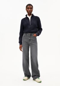 Eine Frau steht da und trägt einen dunkelblauen gerippten Pullover mit halbem Reißverschluss über einem weißen Shirt, weite graue Jeans und weiße Sneaker mit grünen Akzenten.