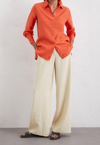 Orange linne knäppskjorta med lång ärm och bröstficka, kombinerad med vida krämfärgade byxor och svarta sandaler, som visar en avslappnad passform.