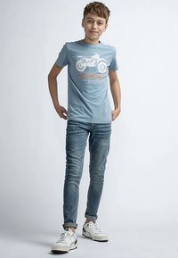 Camiseta de algodón azul claro con un gráfico de motocicleta en blanco y naranja, combinada con vaqueros azules desgastados y zapatillas blancas.