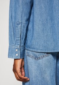 Denim shirt in een blauwtint, met knoopsluiting aan de manchetten met twee zilveren knopen en een relaxed fit. Hand rustend aan de zijkant, waarbij de textuur wordt getoond.