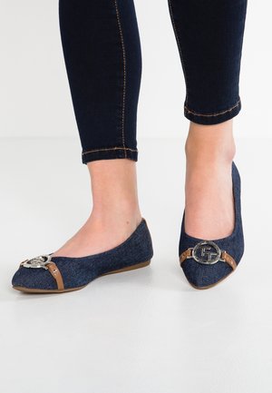 Pieds portant des ballerines en jean bleu foncé avec des bandes marron et des boucles circulaires argentées, assortis à un jean noir ajusté.
