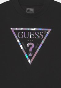 Camiseta de algodón negra con un gran logo triangular holográfico en el centro frente, con el texto "GUESS", "U.S.A." y "WASHED JEANS".