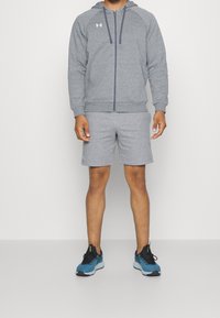 Graue Sportjacke mit Frontreißverschluss, passende Shorts und blauen Laufschuhen. Die Jacke hat ein Logo auf der linken Brust.