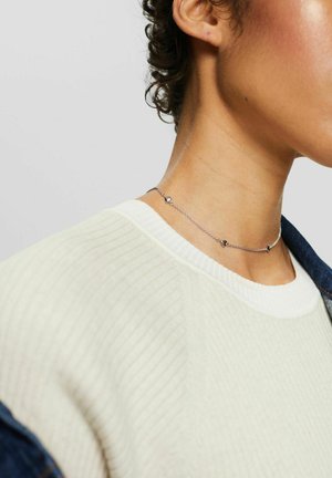 Collana choker in argento con piccoli accenti geometrici, indossata con un maglione bianco a coste. Texture liscia, design della catena delicato.
