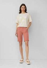 T-shirt beige clair avec un graphique de palmier et de soleil, associé à un short corail à manches retroussées. Le mannequin porte des sandales compensées crème.