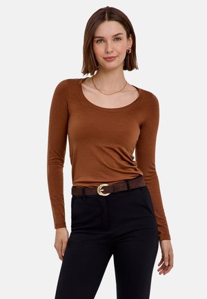 Femme aux cheveux courts et bruns portant un haut marron à manches longues, un pantalon noir et une ceinture marron foncé avec une boucle dorée, debout sur un fond uni.