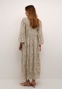 Maxi dress with a smocked back, featuring a swirling beige and brown pattern. The long sleeves are loose and flowing; paired with light-colored shoes.