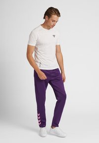 Witte sport t-shirt met een klein logo, gecombineerd met paarse joggers met roze chevronstrepen op het linkerpijp, en witte sneakers.