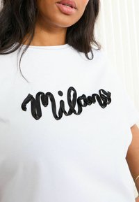 T-shirt blanc avec un texte script en noir brillant "Milano" sur le devant. Le matériau est doux, avec une coupe décontractée et des manches courtes.