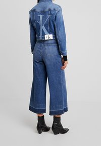 Žena v vykrojených širokých modrých džínách a odpovídající džínsové bundě s krátkým rukávem, v černých kotníkových botách, stojící na jednobarevném pozadí.