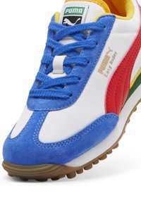 Adidașii PUMA Easy Rider cu un upper alb, accente de piele întoarsă albastră, dungi laterale roșii și detalii multicolore, având o talpă din cauciuc texturat.