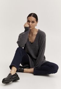 Sivkasti prevelik pulover z V-izrezom in rebrastim vzorcem, v kombinaciji s temno modrimi trenirkami in črnimi loafers. Model sedi na nevtralni podlagi.