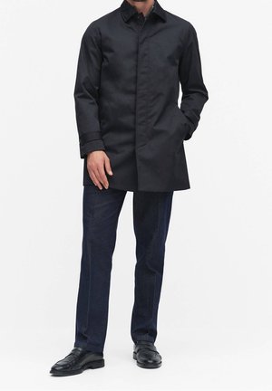Homme portant un manteau noir mi-long, un pantalon bleu foncé et des chaussures habillées noires, debout avec une main dans la poche sur fond blanc.