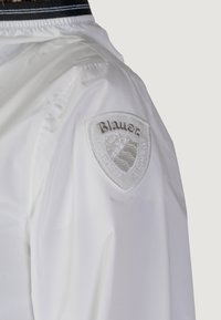 Giacca bianca caratterizzata da un tessuto testurizzato con colletto nero e una patch del logo ricamata in bianco sulla spalla sinistra.