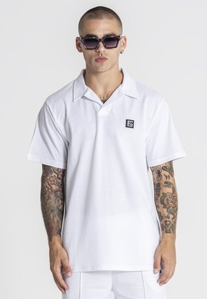 Mand iført hvid poloshirt med krave, solbriller og tatoveringer på begge arme mod en ensartet hvid baggrund.