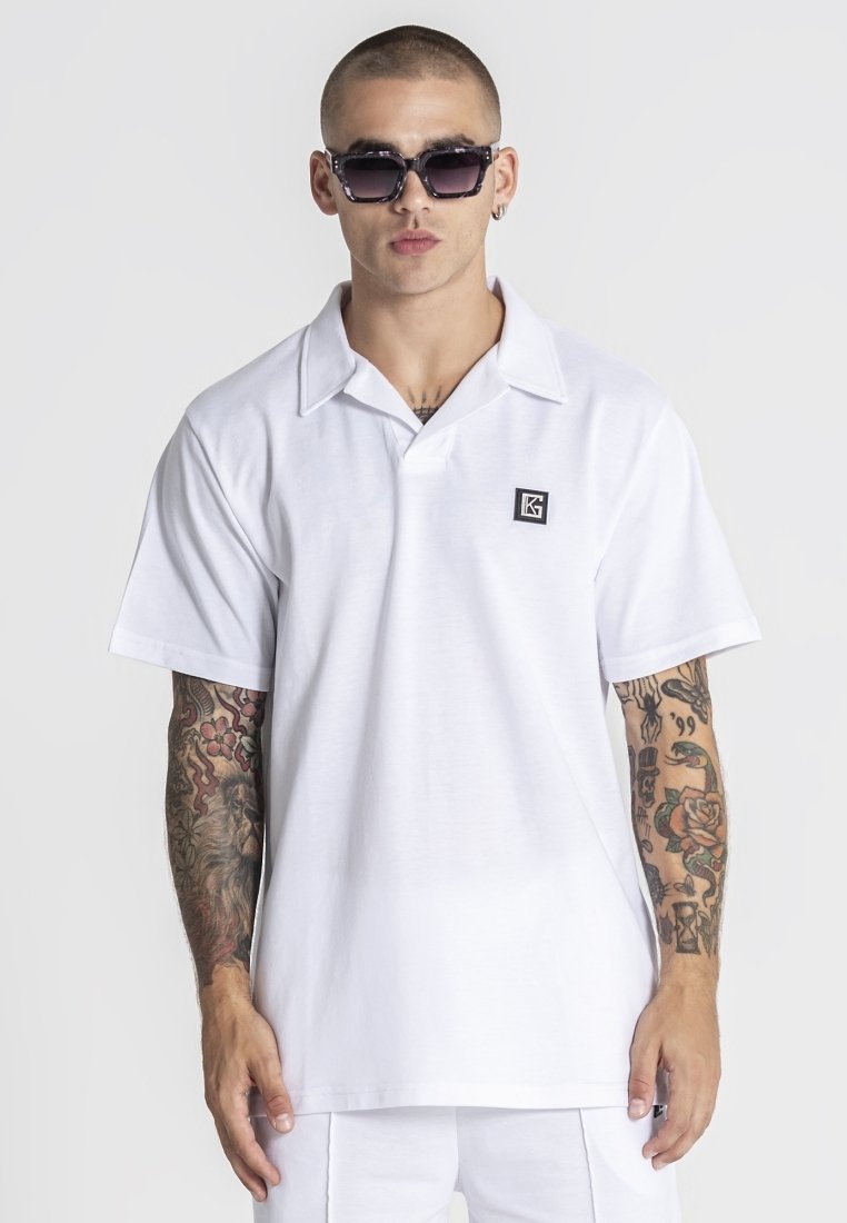 Hombre con camiseta polo blanca con cuello, gafas de sol y tatuajes en ambos brazos sobre un fondo blanco liso.