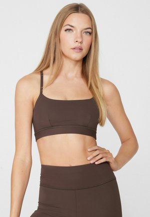 Junge Frau mit langen blonden Haaren, die einen dunkelbraunen Sport-BH und passende hoch taillierte Leggings vor einem schlichten weißen Hintergrund trägt.