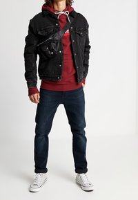 Giacca di jeans nera sopra un hoodie bordeaux, abbinata a jeans scuri e sneaker bianche. Una borsa a tracolla nera è indossata sulla spalla.