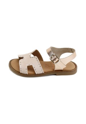 Sandalia plana beige con puntera abierta, parte superior cosida, tira en el tobillo y hebilla dorada sobre suela marrón.