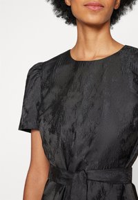 Femme portant une robe courte noire texturée avec un col rond et une ceinture nouée à la taille.