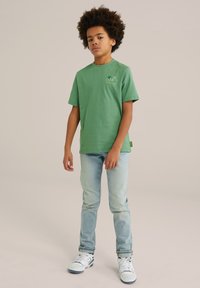 Žalia medvilninė marškin�ė su trumpomis rankovėmis, su maža grafika ant krūtinės. Sujungtos su šviesiai mėlynomis džinsais ir baltais sportbačiais.