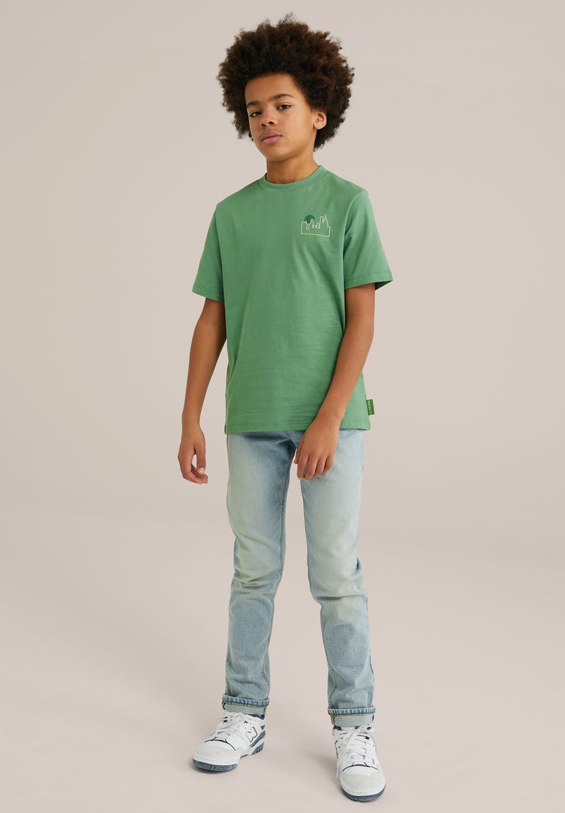 Žalia medvilninė marškinė su trumpomis rankovėmis, su maža grafika ant krūtinės. Sujungtos su šviesiai mėlynomis džinsais ir baltais sportbačiais.