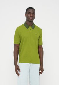 Zöld piké póló kontrasztos sötétzöld gallérral, rövid ujjú, finoman bordázott textúrával. A mellrészen logóval ellátva.