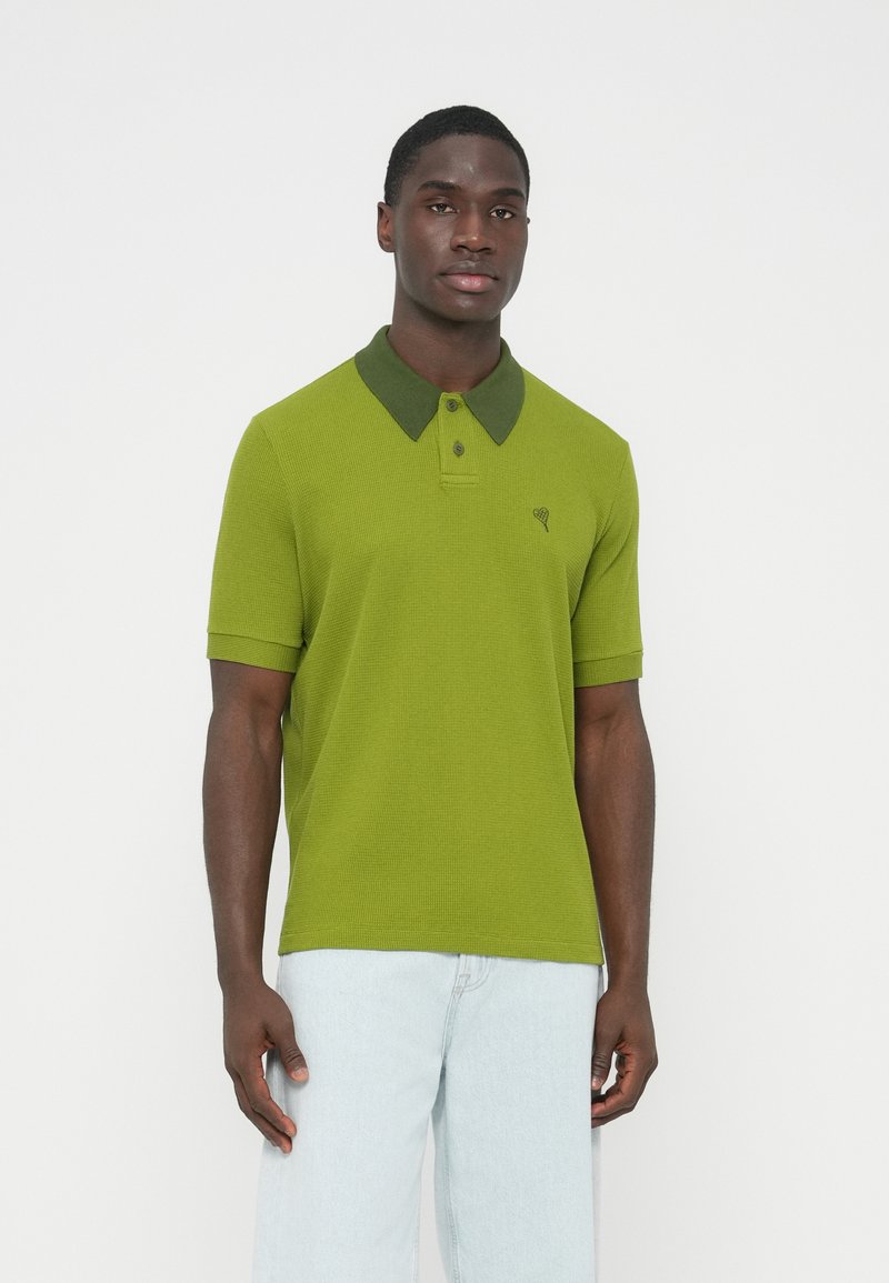 Zöld piké póló kontrasztos sötétzöld gallérral, rövid ujjú, finoman bordázott textúrával. A mellrészen logóval ellátva.