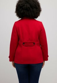 Cappotto rosso in lana con un design strutturato, bottoni posteriori e cuciture sulle spalle. Maniche lunghe e silhouette aderente con cuciture verticali.