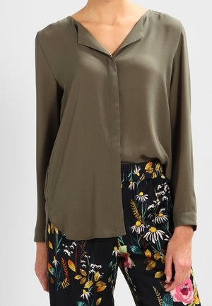 Langarmige olijfgroene blouse gecombineerd met zwarte broek met kleurrijke bloemenpatronen, gedragen door een staande persoon tegen een effen achtergrond.