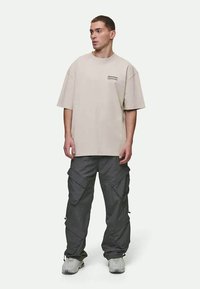 T-shirt oversized bege com uma pequena grafic na parte do peito, combinada com calças cargo cinzas escuras com múltiplos bolsos e zíperes.