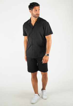 Chemise noire à manches courtes avec col et fermeture à boutons, associée à un short noir. Les accessoires comprennent une montre en argent et des baskets blanches.