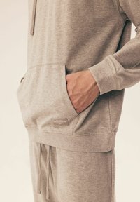 Szara bluza z kapturem, wykonana z miękkiego materiału, z przednią kieszenią, ściągaczami przy rękawach oraz kapturem ze sznurkiem. Posiada małą metkę na kieszeni.