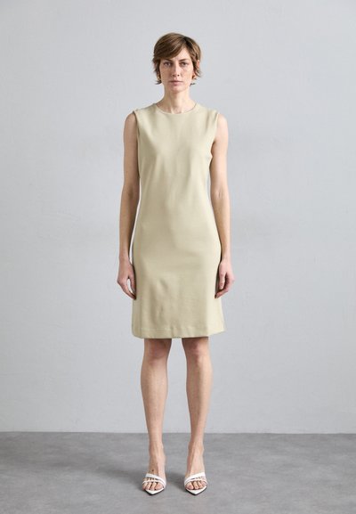 Beige mouwloze jurk met een eenvoudig ontwerp, rechte silhouet en een zachte textuur. Gecombineerd met witte hakken met bandjes.