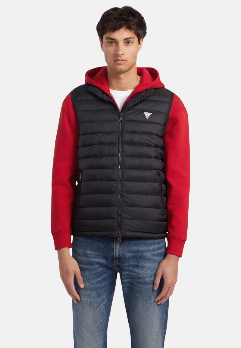 Chaleco negro acolchado tipo puffer con cremallera y patrón acolchado horizontal. Lleva una sudadera con capucha roja debajo. Destaca un logotipo triangular en el pecho izquierdo.
