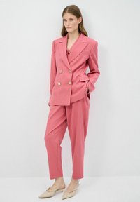 Vaaleanpunainen kaksirivinen bleiseri, jossa on kultanapit, yhdistettynä siihen sopiviin housuihin. Kangas on sileä, ja kengät ovat beigejä, joissa on terävä kärki.