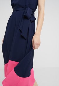 Mörkblå klänning med en rosa fåll, med ett knytsmitt och mjukt tyg. Designen inkluderar en flödande silhuett och texturerad detalj.