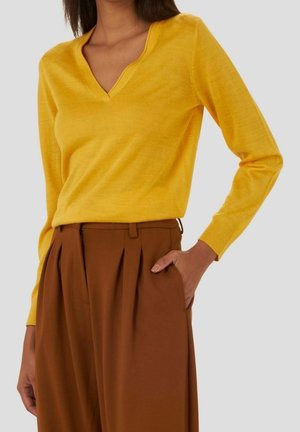 Mujer con suéter amarillo brillante de manga larga y escote en V, metido dentro de pantalones marrones de pierna ancha y cintura alta, con la mano en el bolsillo.