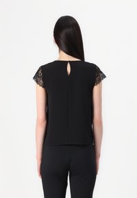 Sort bluse med blonde cap-sleeves, detaljer med nøglehul i ryggen, glat tekstur og enkelt design. Sammen med mørke bukser i en minimalistisk stil.