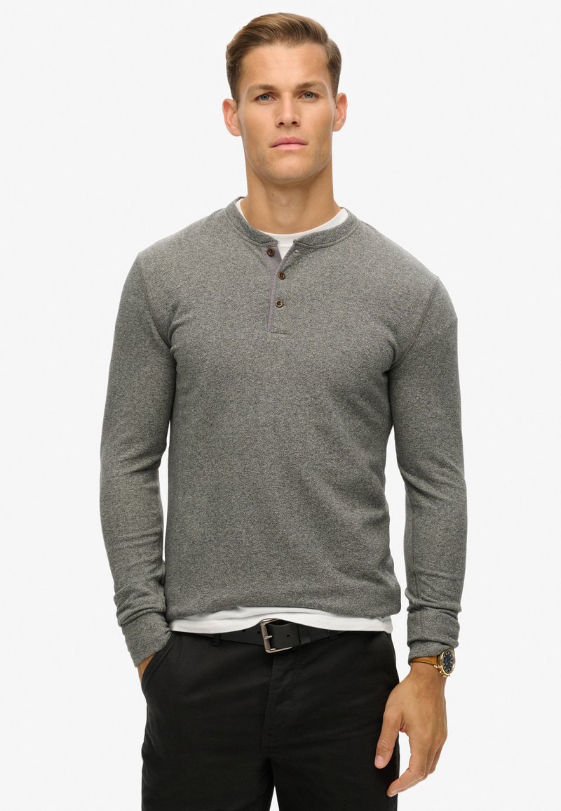 Graues Langarm-Henley-Shirt aus strukturiertem Stoff, mit drei braunen Knöpfen und rundem Ausschnitt, kombiniert mit schwarzen Hosen und einer Uhr.